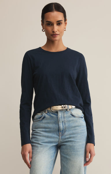 A woman wearing a navy long-sleeved t-shirt with rib detailing at the neck and a round neckline, paired with light blue jeans. She is also wearing a belt with a metallic buckle.