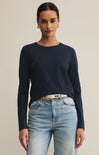 A woman wearing a navy long-sleeved t-shirt with rib detailing at the neck and a round neckline, paired with light blue jeans. She is also wearing a belt with a metallic buckle.