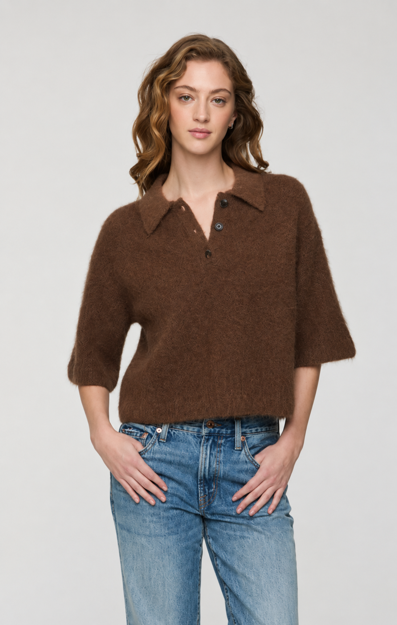 A woman modeling a brown fuzzy knit polo sweater with three-quarter sleeves and a buttoned collar, paired with blue jeans, against a light gray studio background.