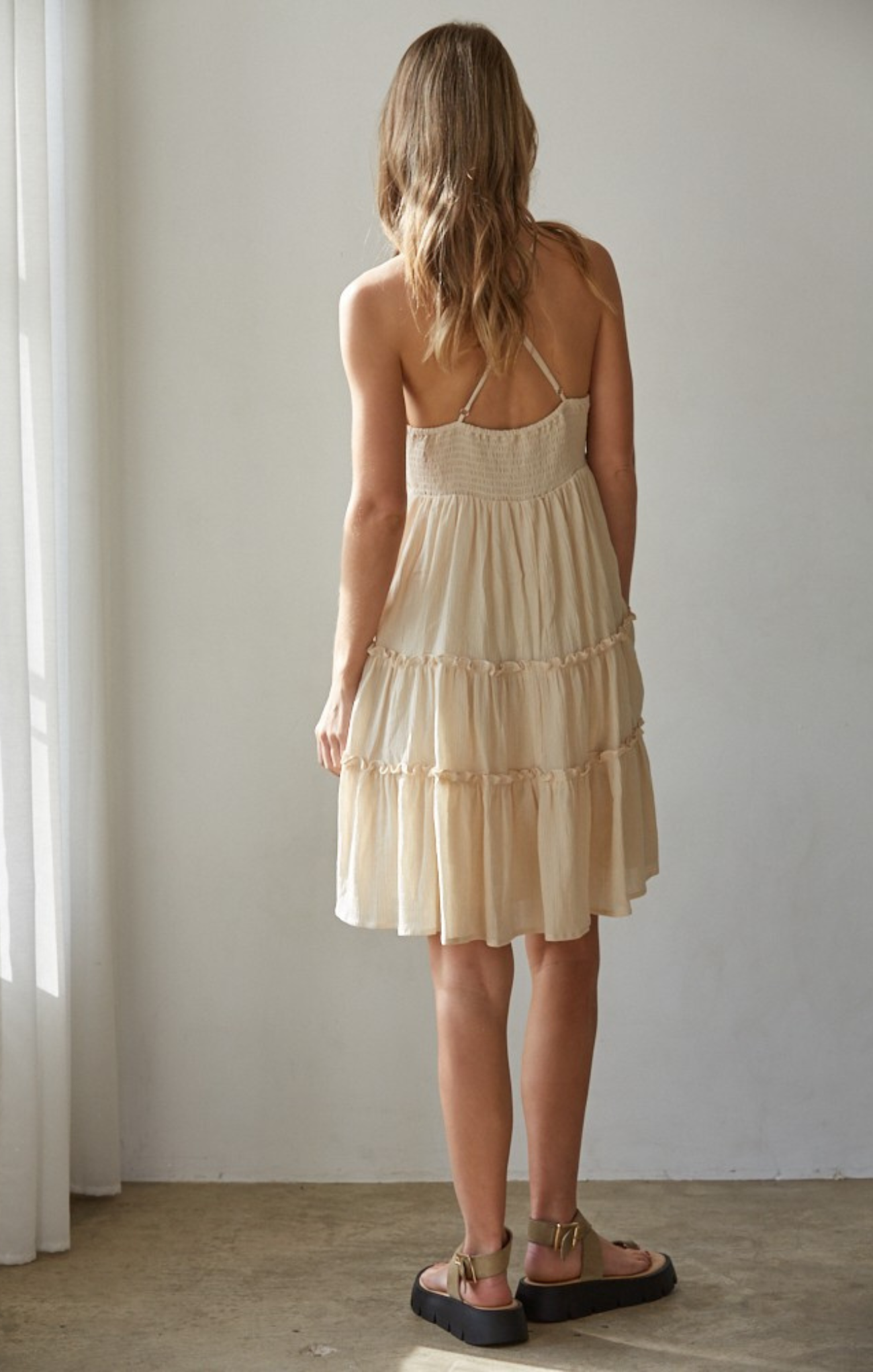 Woman wearing a beige tiered dress standing against a plain wall.