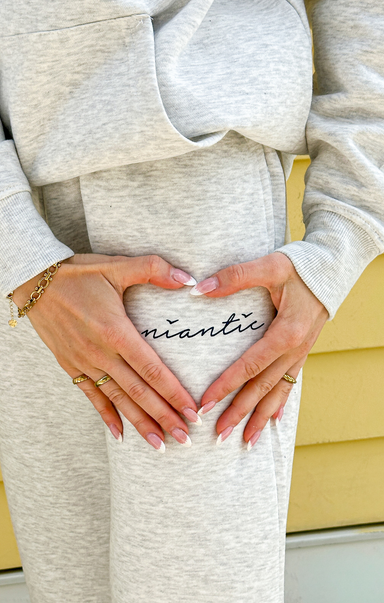 Pregnant person wearing a gray outfit with 'anticipating' written on it, holding hands in a heart shape.