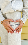 Pregnant person wearing a gray outfit with 'anticipating' written on it, holding hands in a heart shape.