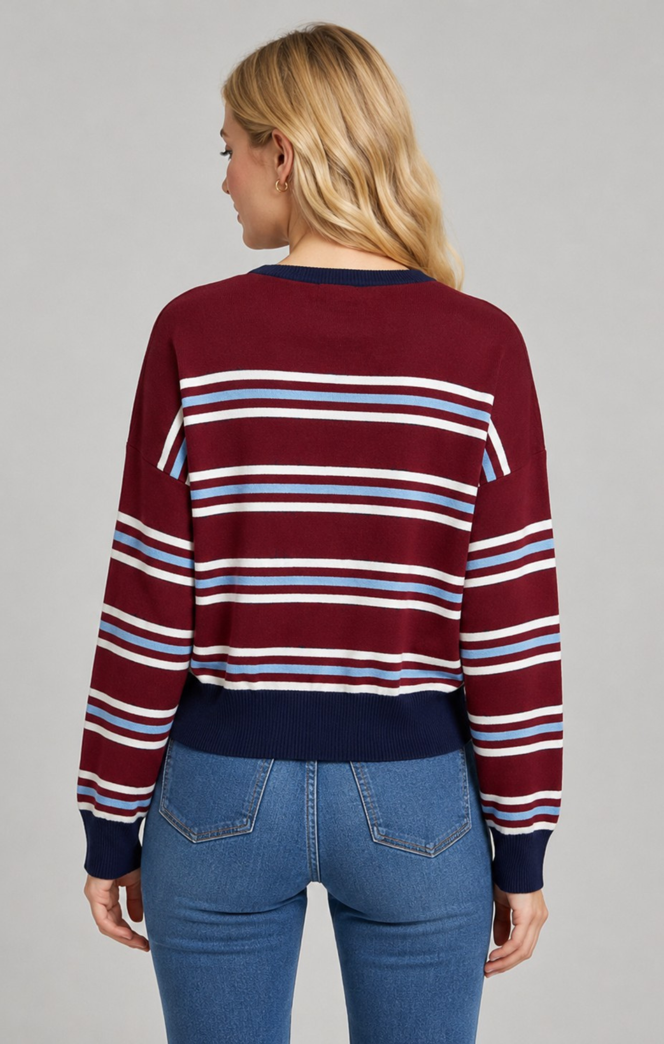 A woman viewed from the back wearing a burgundy long-sleeve knit sweater with horizontal navy, white, and light blue stripes, paired with blue jeans, against a solid gray studio background.
