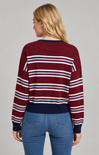 A woman viewed from the back wearing a burgundy long-sleeve knit sweater with horizontal navy, white, and light blue stripes, paired with blue jeans, against a solid gray studio background.