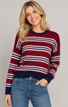 A woman wearing a burgundy, navy, white, and light blue striped crewneck sweater with blue jeans, photographed against a solid light gray studio background.