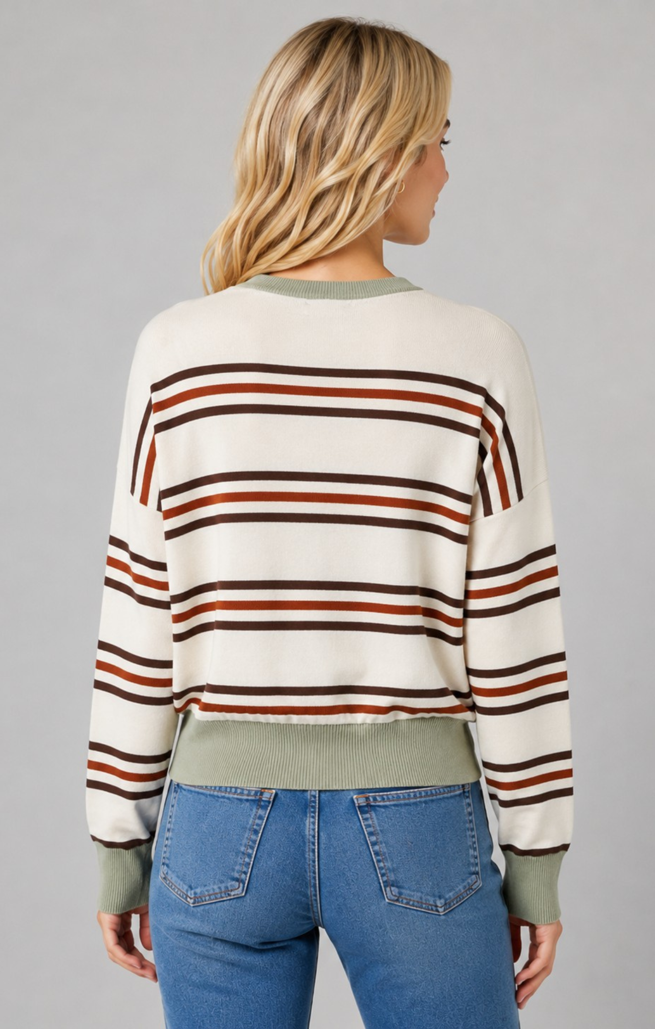 A woman viewed from the back wearing a cropped knit sweater with horizontal stripes in rust brown, dark brown, and olive green on an off-white base, paired with blue jeans against a solid gray studio background.