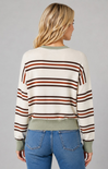 A woman viewed from the back wearing a cropped knit sweater with horizontal stripes in rust brown, dark brown, and olive green on an off-white base, paired with blue jeans against a solid gray studio background.