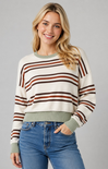 A woman wearing a cream-colored crewneck sweater with brown and rust-orange horizontal stripes, paired with blue jeans, against a solid gray studio background.