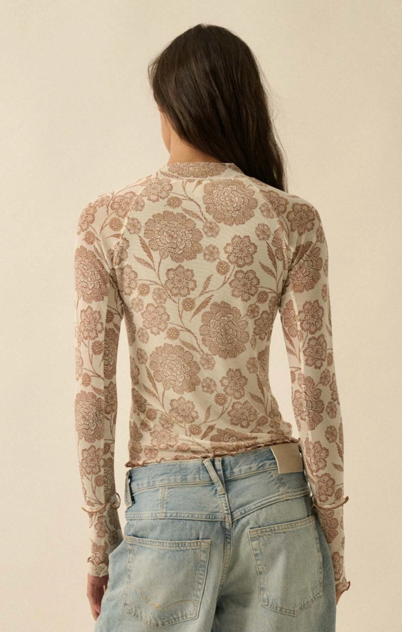 A woman viewed from the back wearing a fitted long-sleeve top with an allover floral pattern in warm beige and brown tones, paired with light blue jeans against a plain cream background.