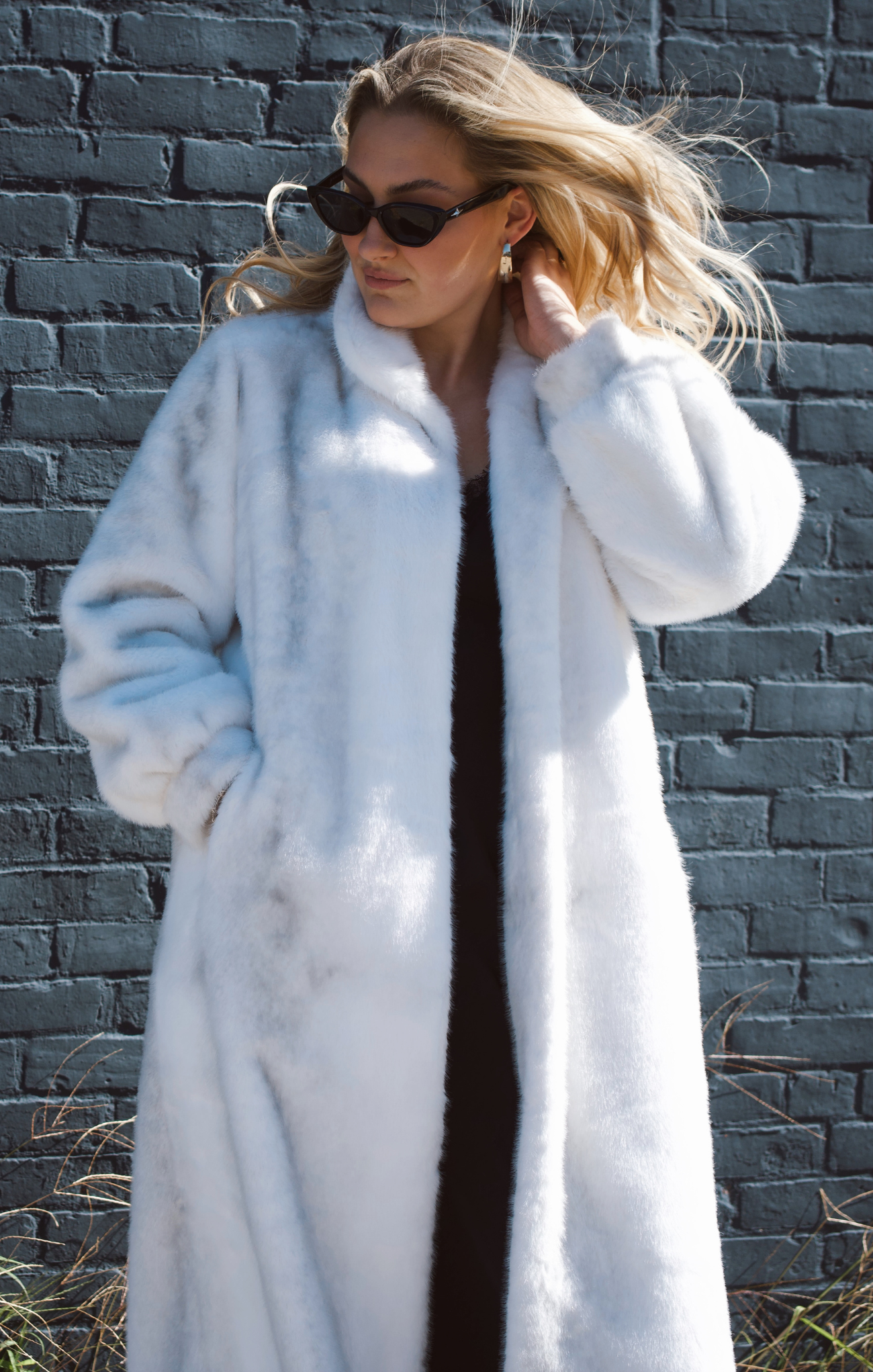 Woman wearing a long white fur coat against a brick wall.
