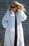 Woman wearing a long white fur coat against a brick wall.