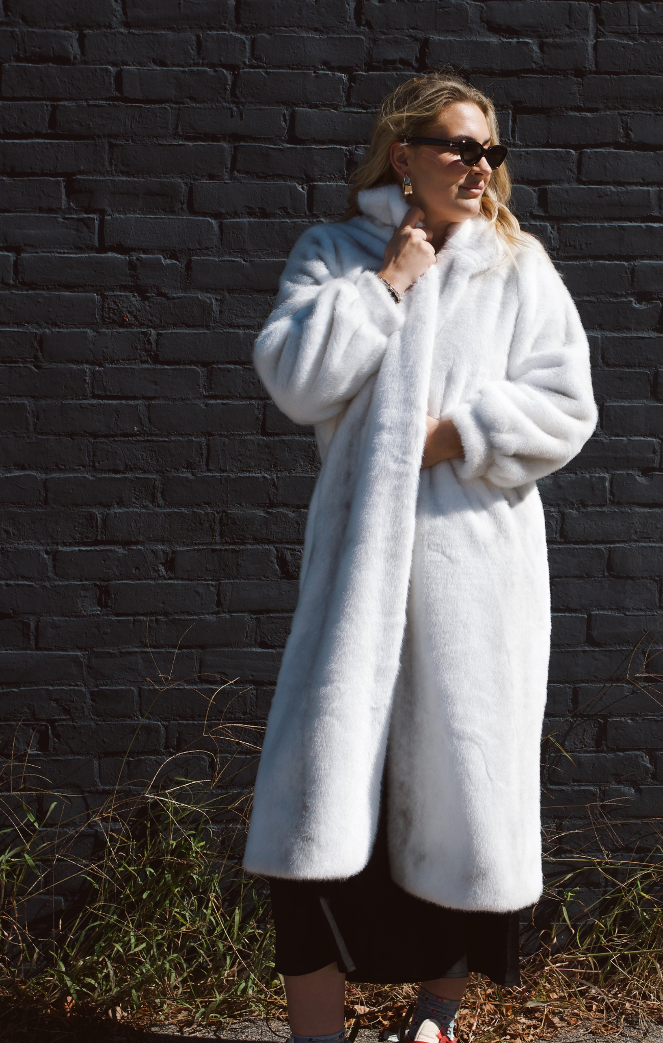 Woman wearing a white fur coat against a dark brick wall.