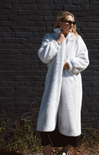 Woman wearing a white fur coat against a dark brick wall.