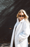 Woman wearing a white fur coat and sunglasses against a dark brick wall.