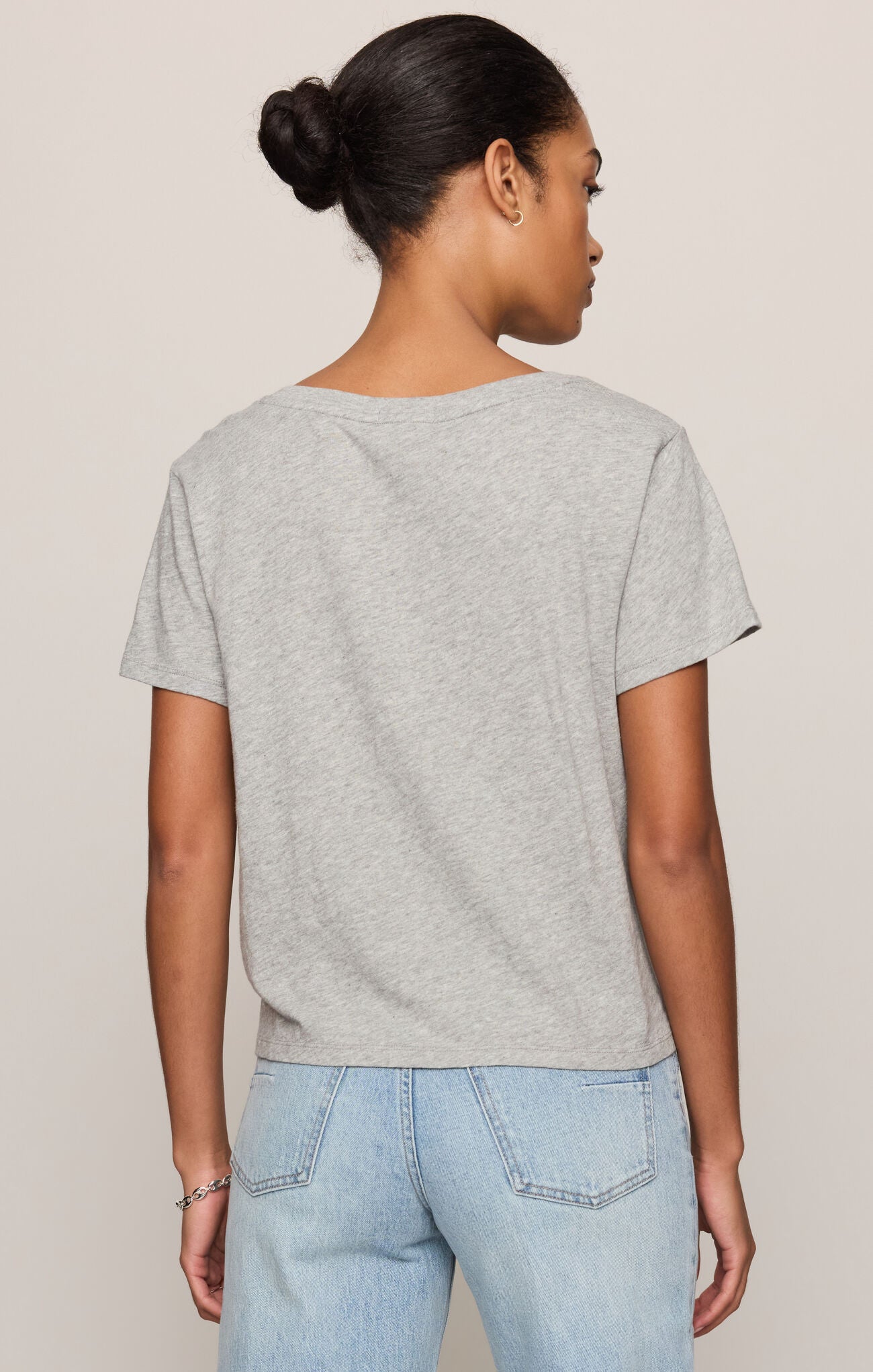 A woman shown from the back wearing a light gray heather short-sleeve t-shirt and light blue jeans, set against a plain beige studio background.