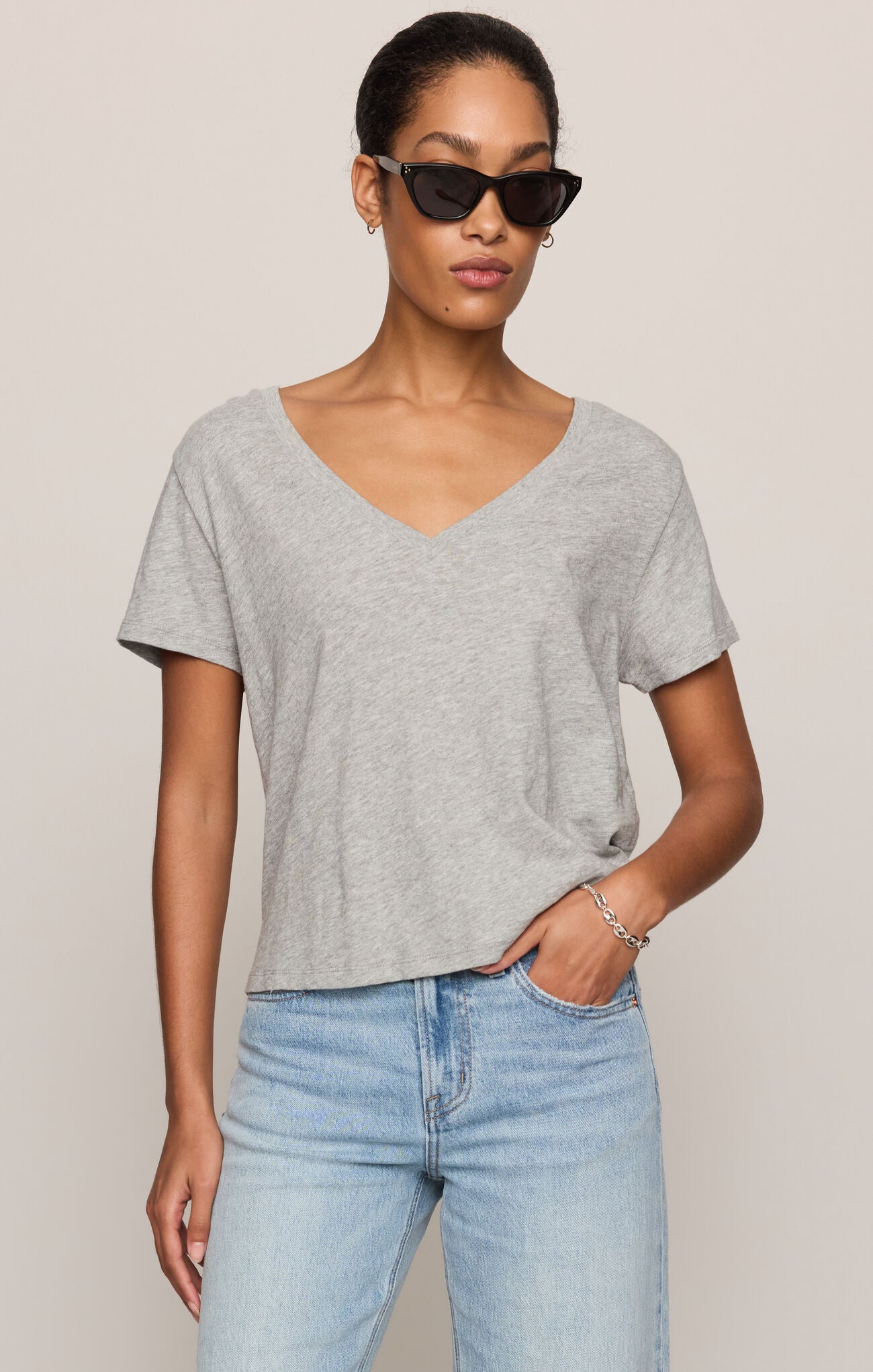 A woman modeling a light gray heathered short-sleeve V-neck t-shirt paired with light-wash blue jeans, photographed against a solid light beige studio background.