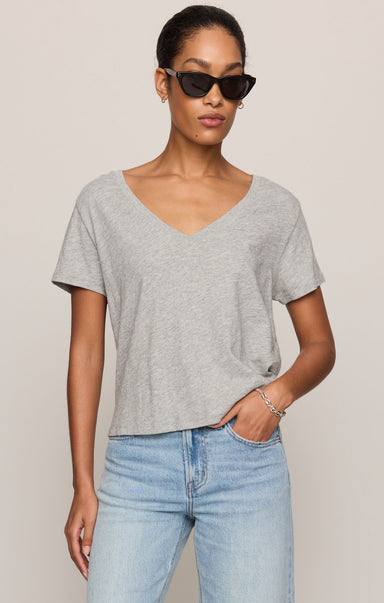 A woman modeling a light gray heathered short-sleeve V-neck t-shirt paired with light-wash blue jeans, photographed against a solid light beige studio background.