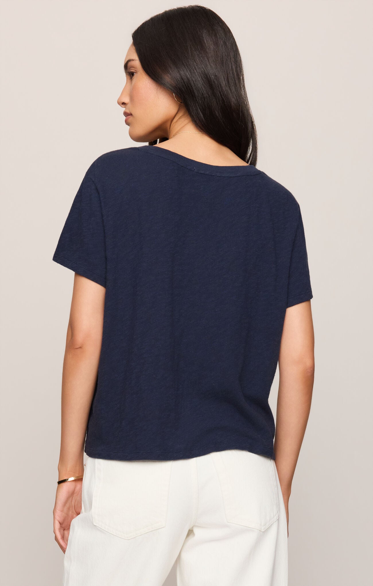 A woman shown from the back wearing a short-sleeve navy blue t-shirt and white pants, photographed against a plain light beige studio background.