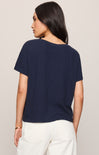 A woman shown from the back wearing a short-sleeve navy blue t-shirt and white pants, photographed against a plain light beige studio background.
