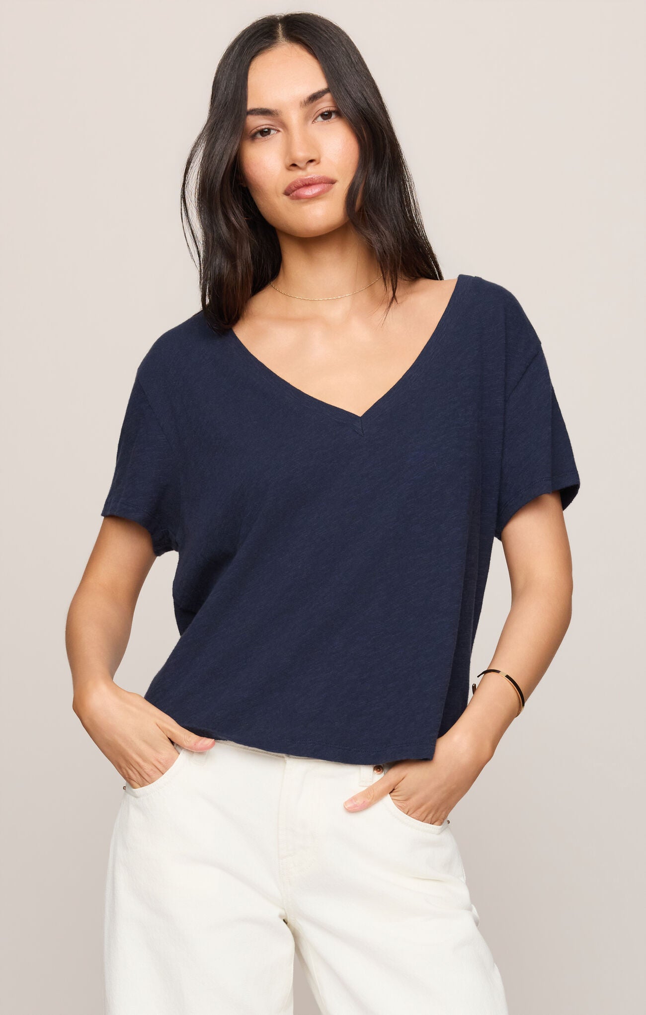 A woman modeling a navy blue short-sleeve V-neck top with white pants, standing against a plain light beige studio background.