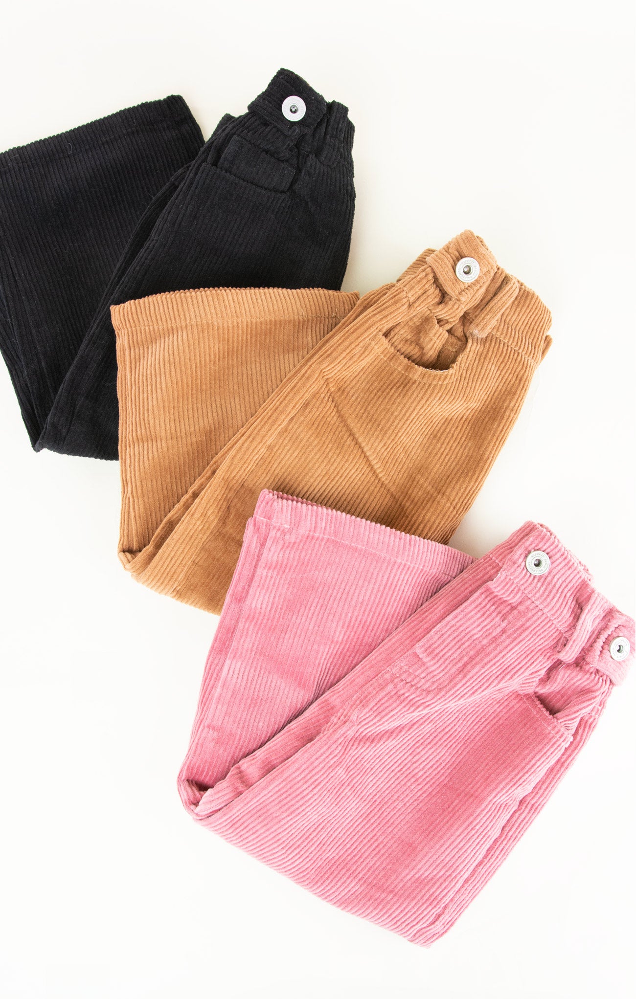 Three pairs of folded corduroy pants in black, brown, and pink on a white background.