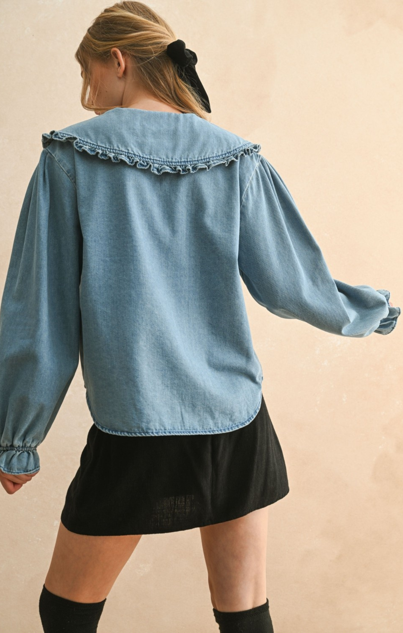Woman wearing a blue denim shirt with ruffled collar and black skirt on a beige background