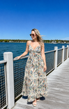 Woman in a floral dress standing on a dock by a body of water.