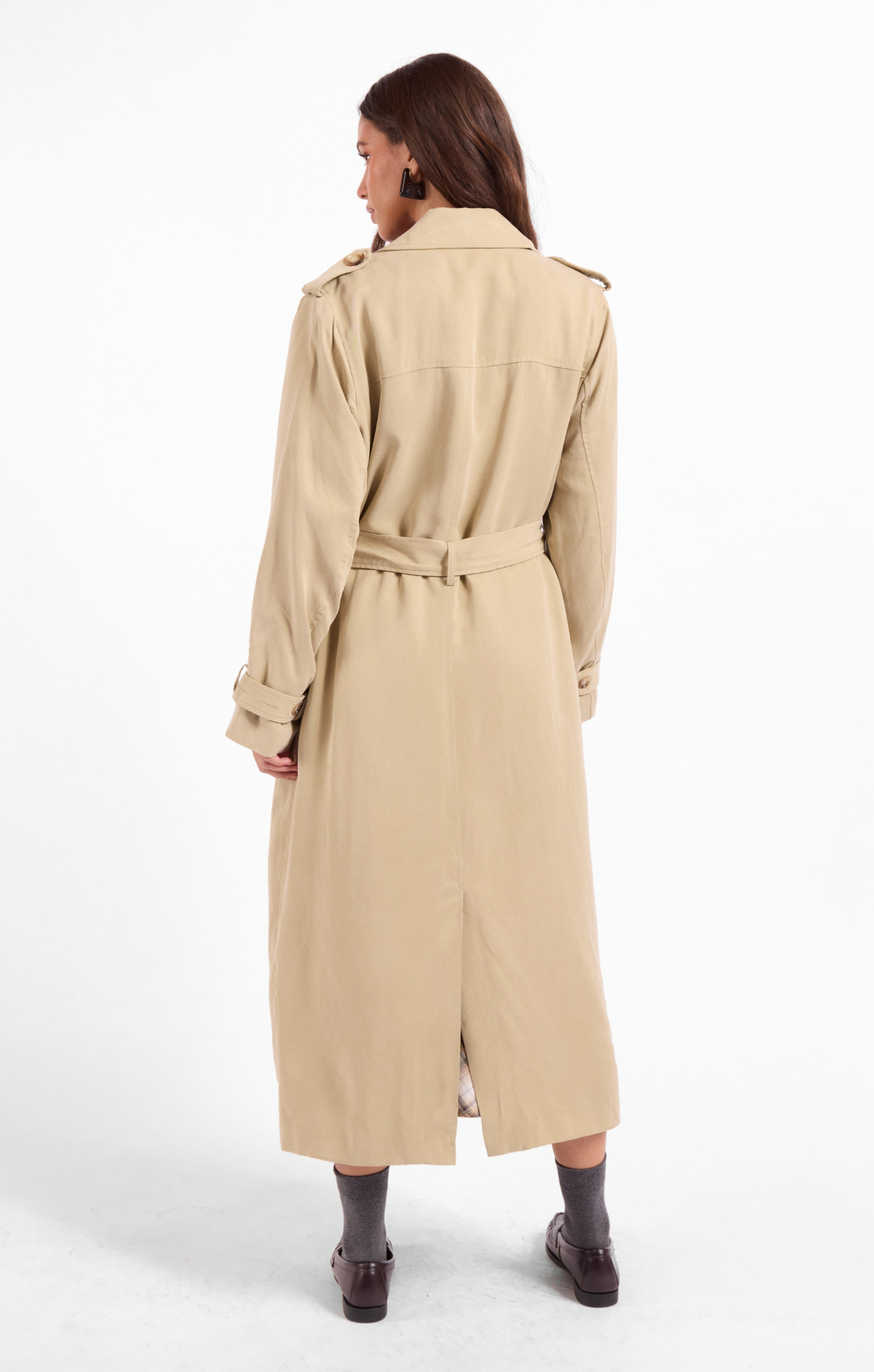 A woman shown from the back wearing a long beige trench coat with a matching waist belt, paired with dark socks and brown shoes, against a plain white studio background.