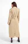 A woman shown from the back wearing a long beige trench coat with a matching waist belt, paired with dark socks and brown shoes, against a plain white studio background.