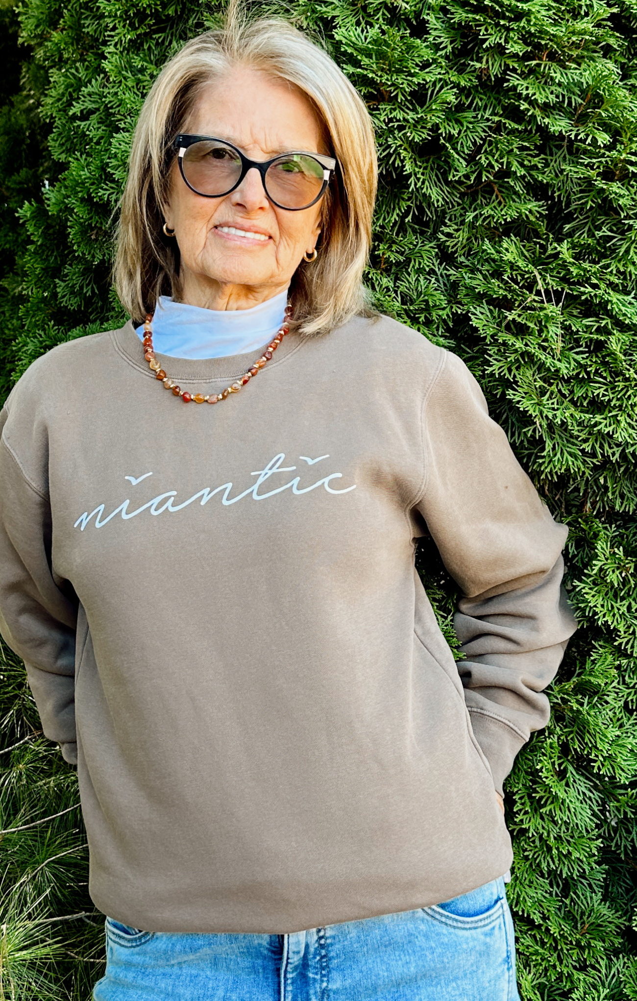 Person wearing a brown crewneck with 'victoria' text, standing against a green hedge background.