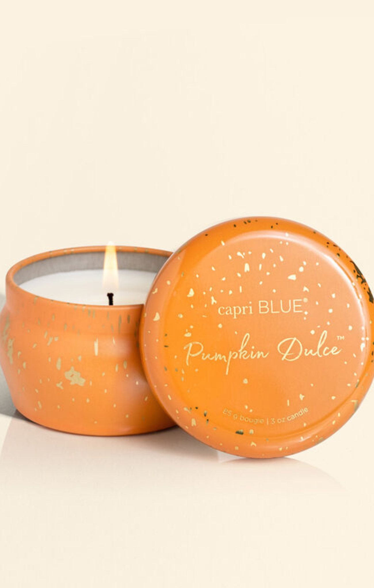 An orange speckled metal candle tin with its lid leaning against it, showing a lit candle inside. The lid is printed with the brand name 'capri BLUE' and the scent 'Pumpkin Dulce'.