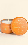 An orange speckled metal candle tin with its lid leaning against it, showing a lit candle inside. The lid is printed with the brand name 'capri BLUE' and the scent 'Pumpkin Dulce'.