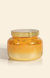 A small clear glass jar filled with golden yellow wax or honey-like substance, topped with a metallic gold lid, photographed on a clean off-white background.