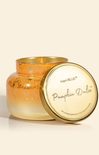A Capri Blue 'Pumpkin Dolce' scented candle in a textured, gold-toned glass jar with its lid leaning against it, set against a warm cream background.