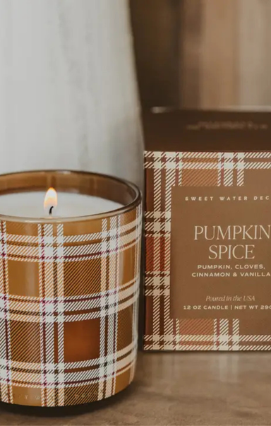 A lit candle in a brown glass jar with a tan and white plaid pattern, shown next to its matching brown box labeled 'Pumpkin Spice' on a wooden surface.