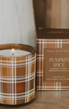 A lit candle in a brown glass jar with a tan and white plaid pattern, shown next to its matching brown box labeled 'Pumpkin Spice' on a wooden surface.