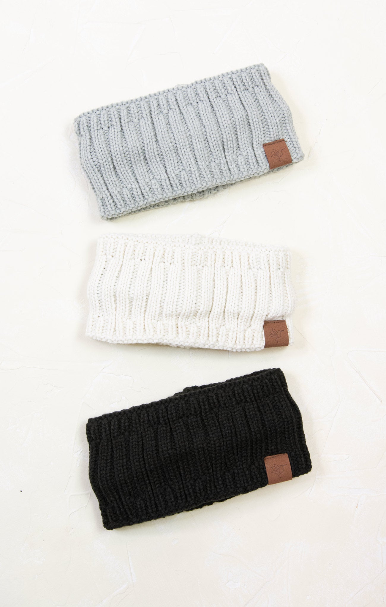Three knitted headbands in gray, white, and black with brown leather patches on a white background.