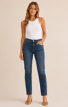 A woman modeling a pair of high-waisted, ankle-length blue jeans with a white sleeveless top and beige heeled sandals, standing against a solid beige studio background.