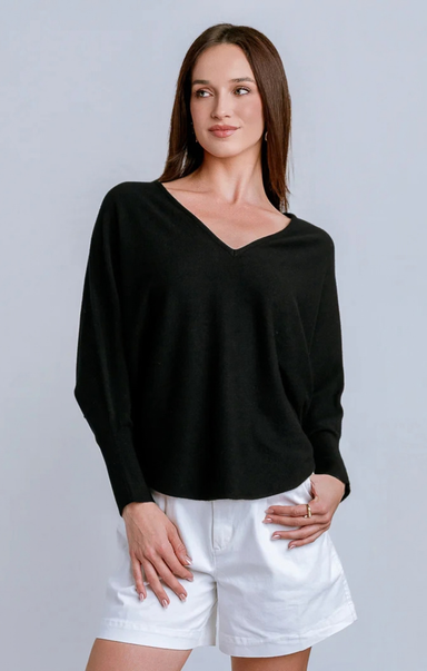 A woman modeling a black long-sleeve V-neck knit top with white shorts against a light gray studio background.