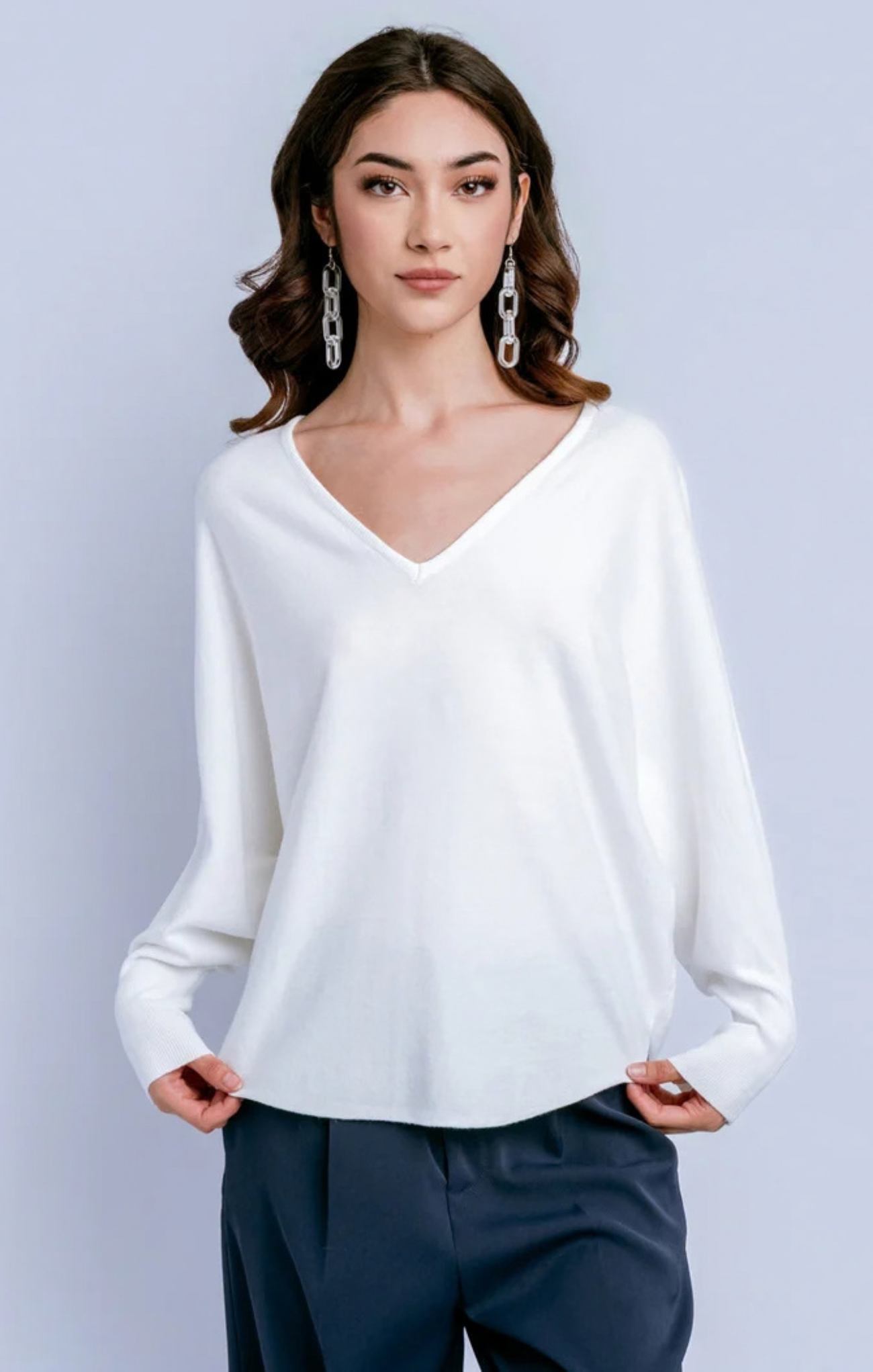 A woman modeling a white long-sleeve V-neck knit top with dark navy pants, standing against a light gray studio background.