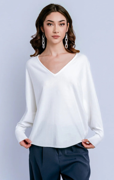 A woman modeling a white long-sleeve V-neck knit top with dark navy pants, standing against a light gray studio background.