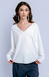 A woman modeling a white long-sleeve V-neck knit top with dark navy pants, standing against a light gray studio background.