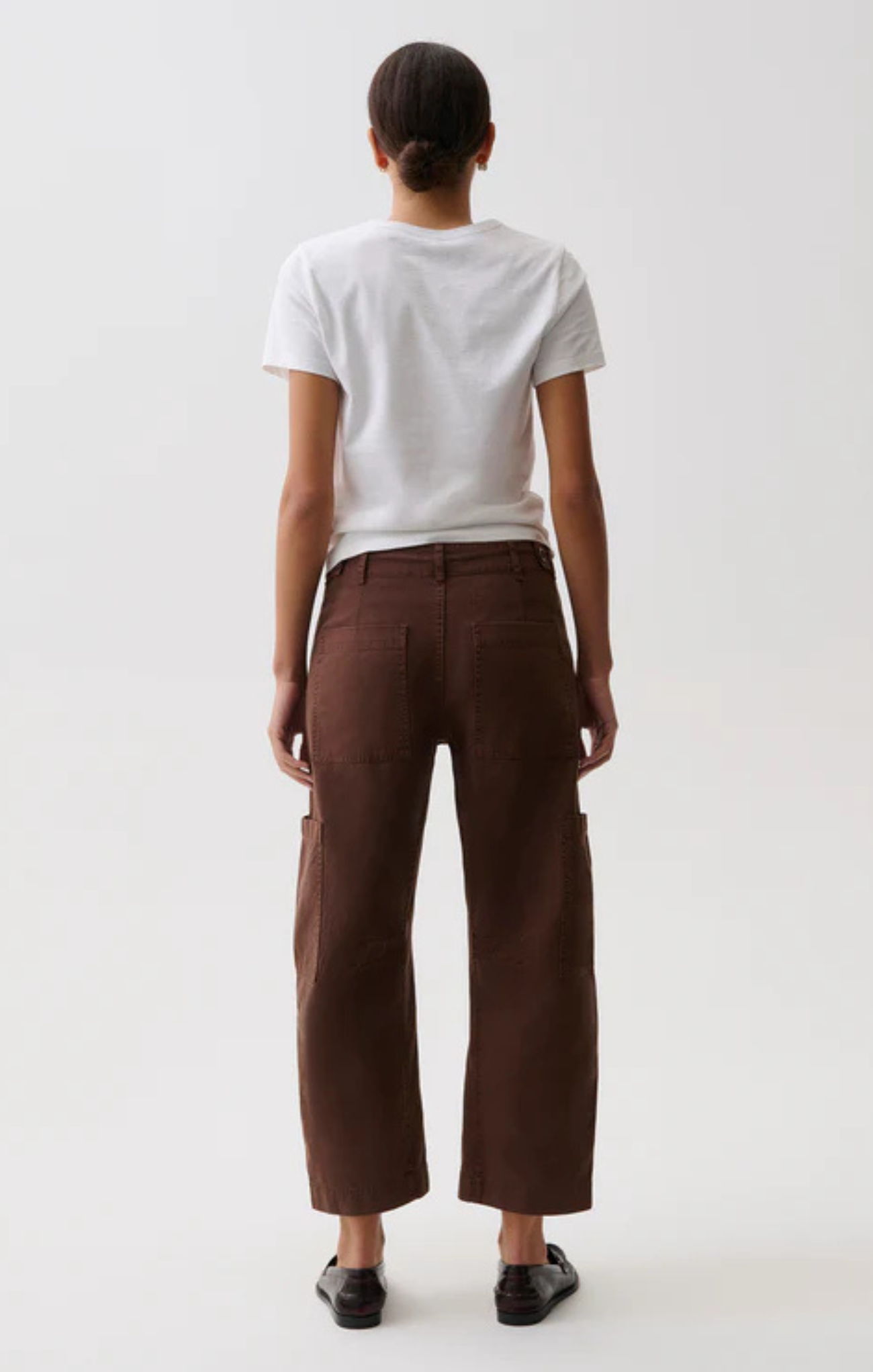 A model shown from the back wearing brown wide-leg cropped trousers, a white t-shirt, and black shoes against a plain light gray background.