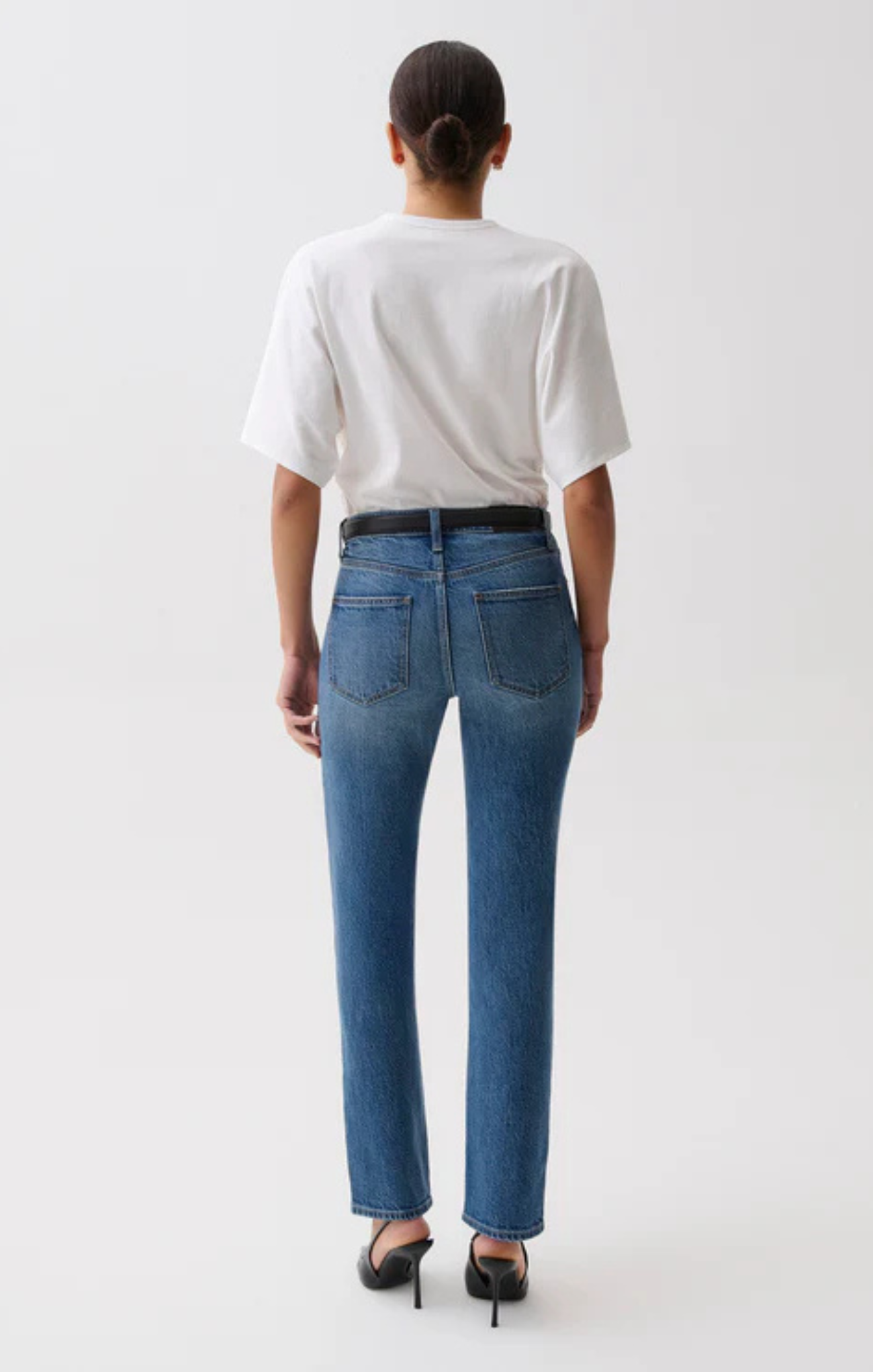 A model shown from the back wearing blue high-waisted straight-leg jeans, a white t-shirt, a black belt, and black heels against a plain white studio background.