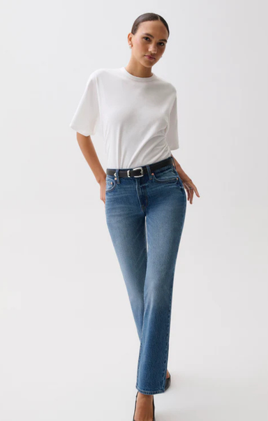 A woman modeling a white short-sleeve crewneck t-shirt and blue straight-leg jeans with a black belt, standing against a plain white studio background.
