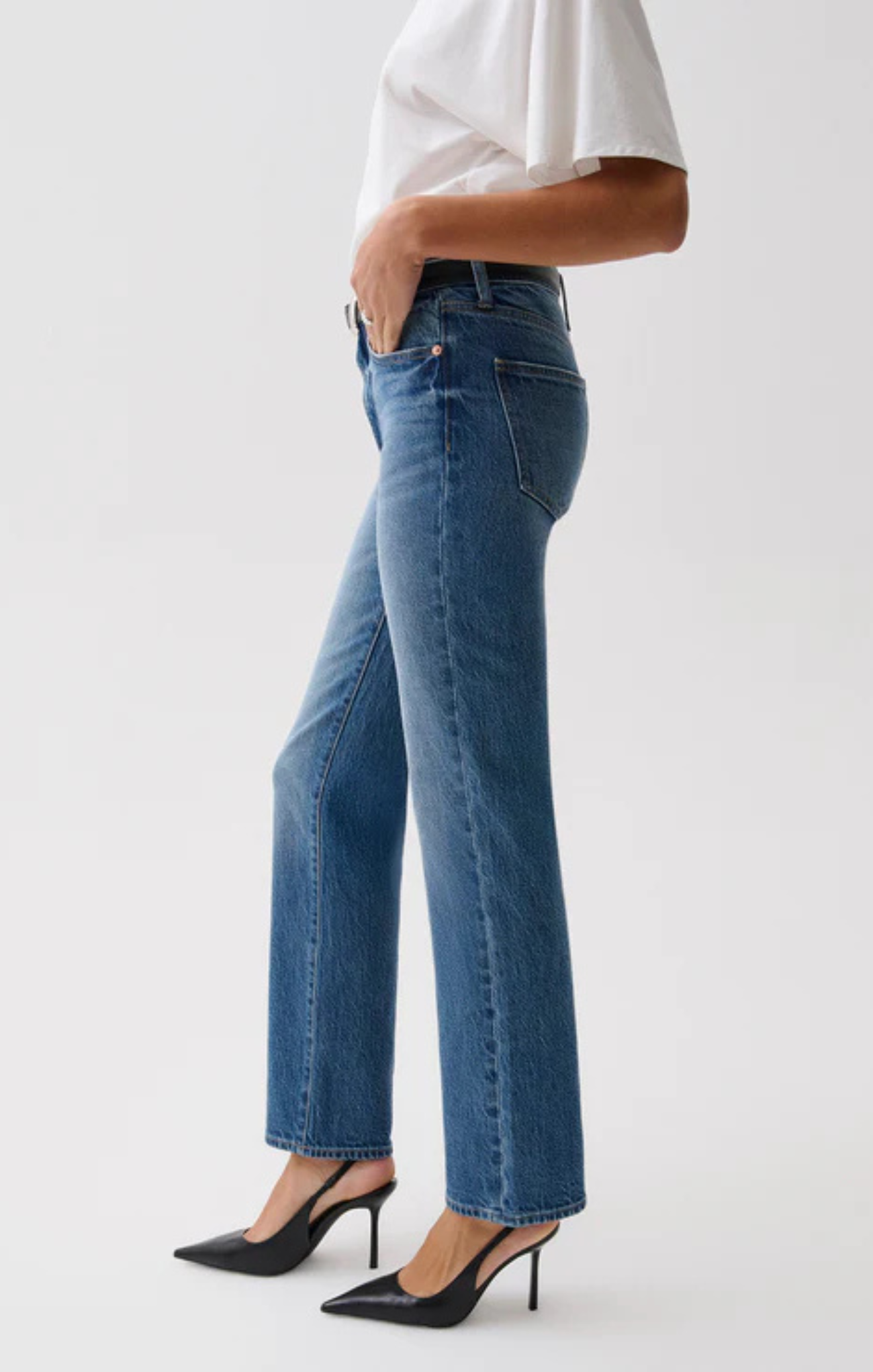 A side-profile view of a model wearing medium-wash blue straight-leg jeans, a white top, and black pointed-toe stiletto heels against a light gray studio background.