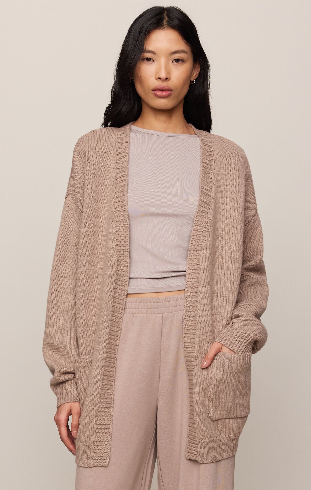 Woman wearing a beige cardigan over a light gray top and beige pants on a plain background