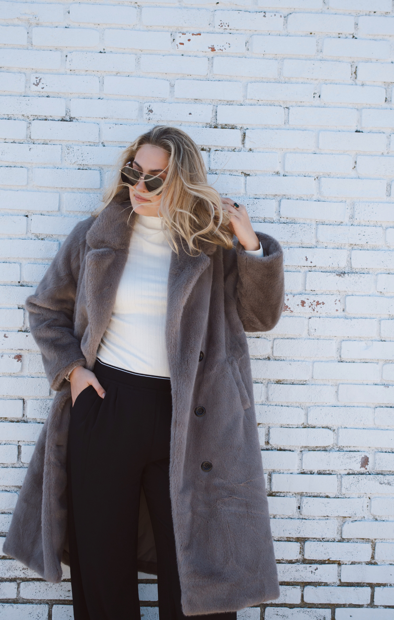 Woman wearing a long gray coat against a white brick wall