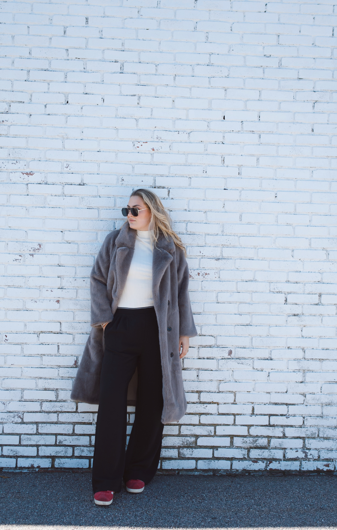 Person wearing a gray coat and sunglasses against a white brick wall