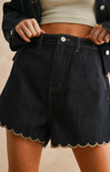 Person wearing black denim shorts with scalloped hem on a neutral background
